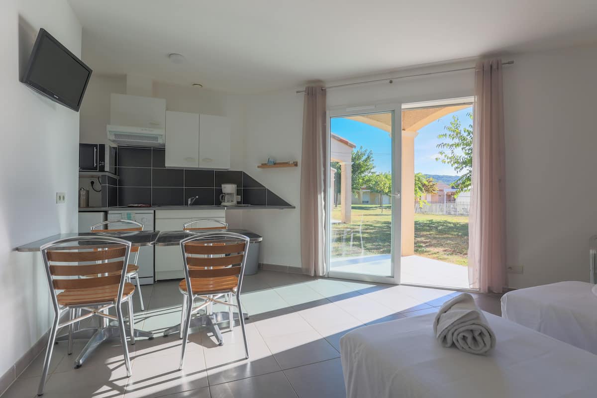 A modern hotel room with a kitchen area, a dining table with chairs, a bed, and a large sliding glass door leading to a balcony with a view of the outdoors.