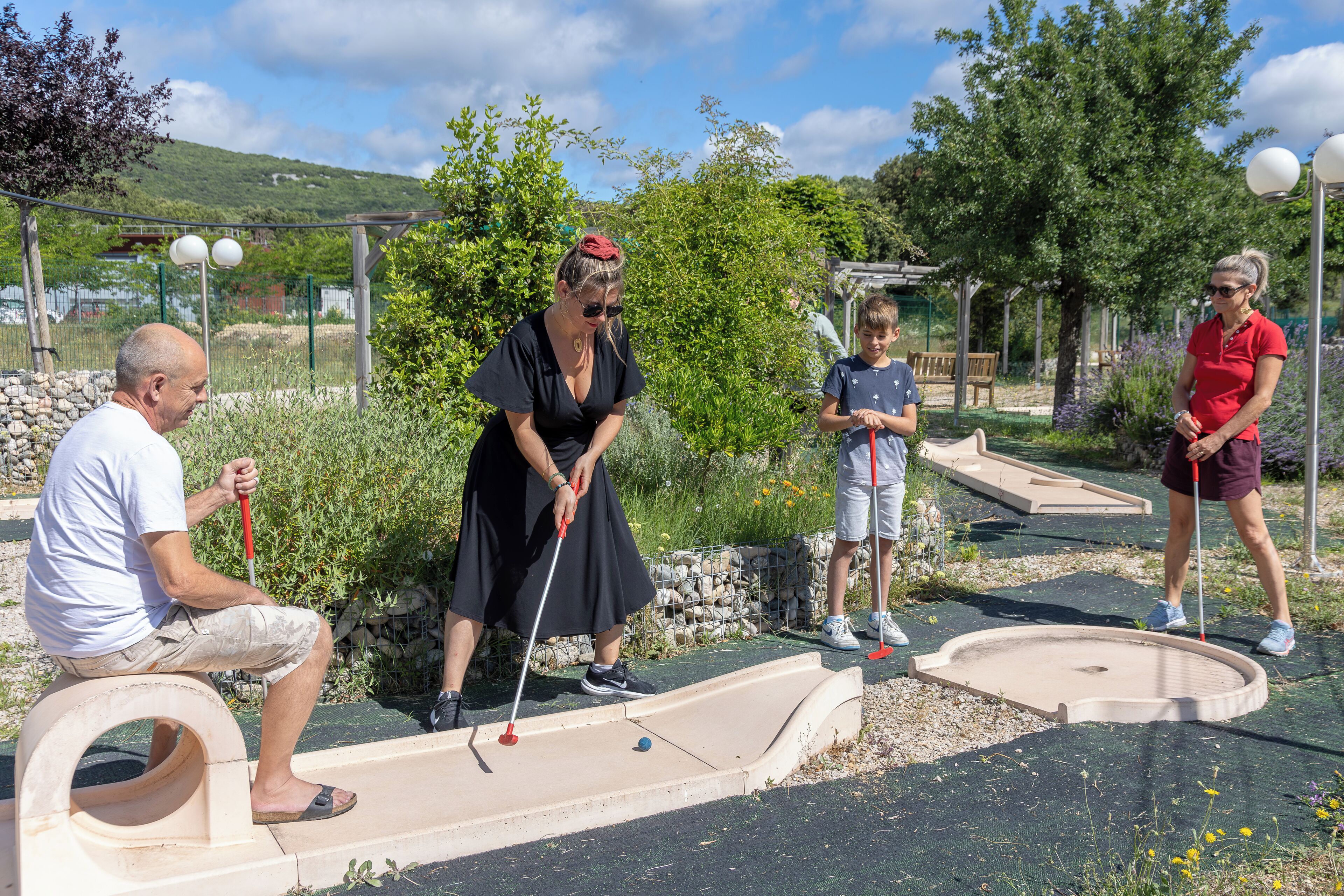 A group of people playing mini-golf in an outdoor setting with a green landscape and trees in the background.