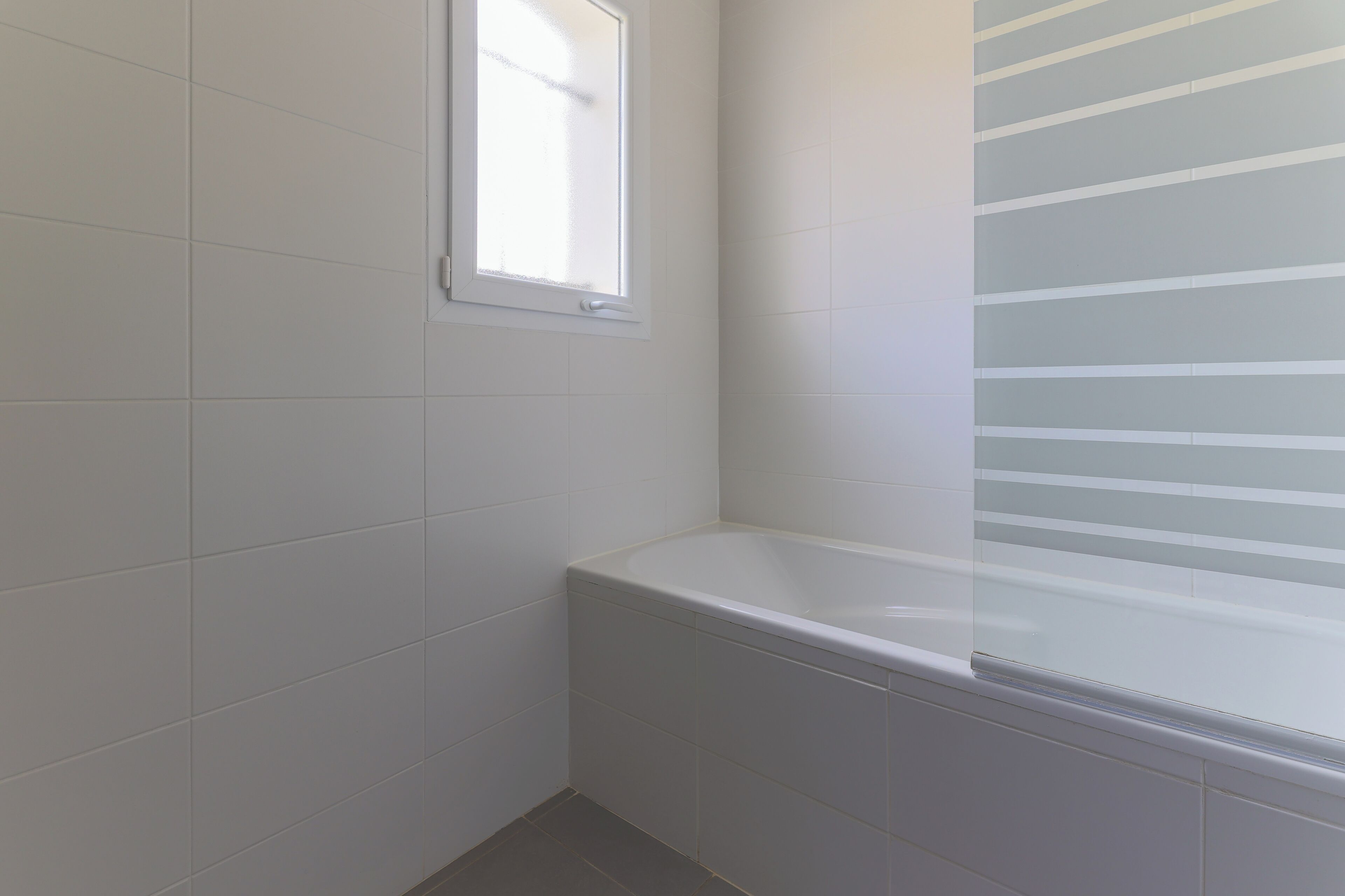 Une salle de bain avec des murs recouverts de carreaux blancs, une fenêtre et une baignoire.