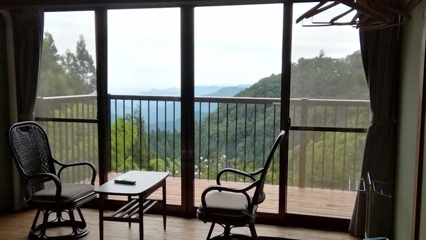 Una habitación con una cama, una mesa con tetera y bandeja, dos sillas y vistas a un balcón con barandilla y vegetación.