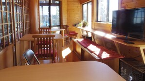 Interior acogedor de una cabaña con paredes de madera, un comedor con sillas y una mesa, un televisor sobre un mueble bajo y una ventana con vistas a árboles.