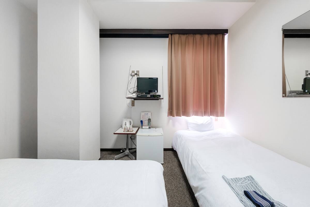 Une chambre d’hôtel avec deux lits, un bureau avec un ordinateur, une petite table avec un téléphone et une fenêtre avec des rideaux.