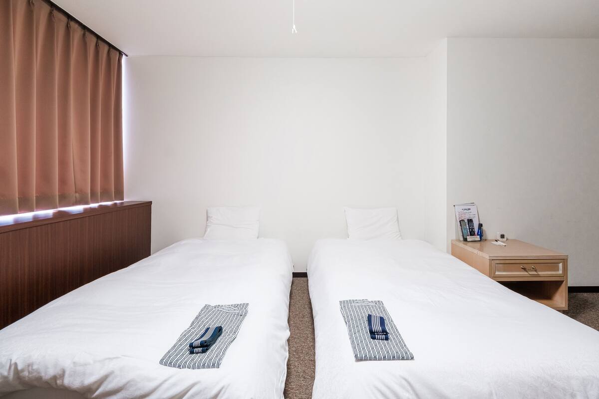Une chambre d’hôtel avec deux lits simples, une table de chevet en bois, un petit tiroir et une fenêtre avec des rideaux.