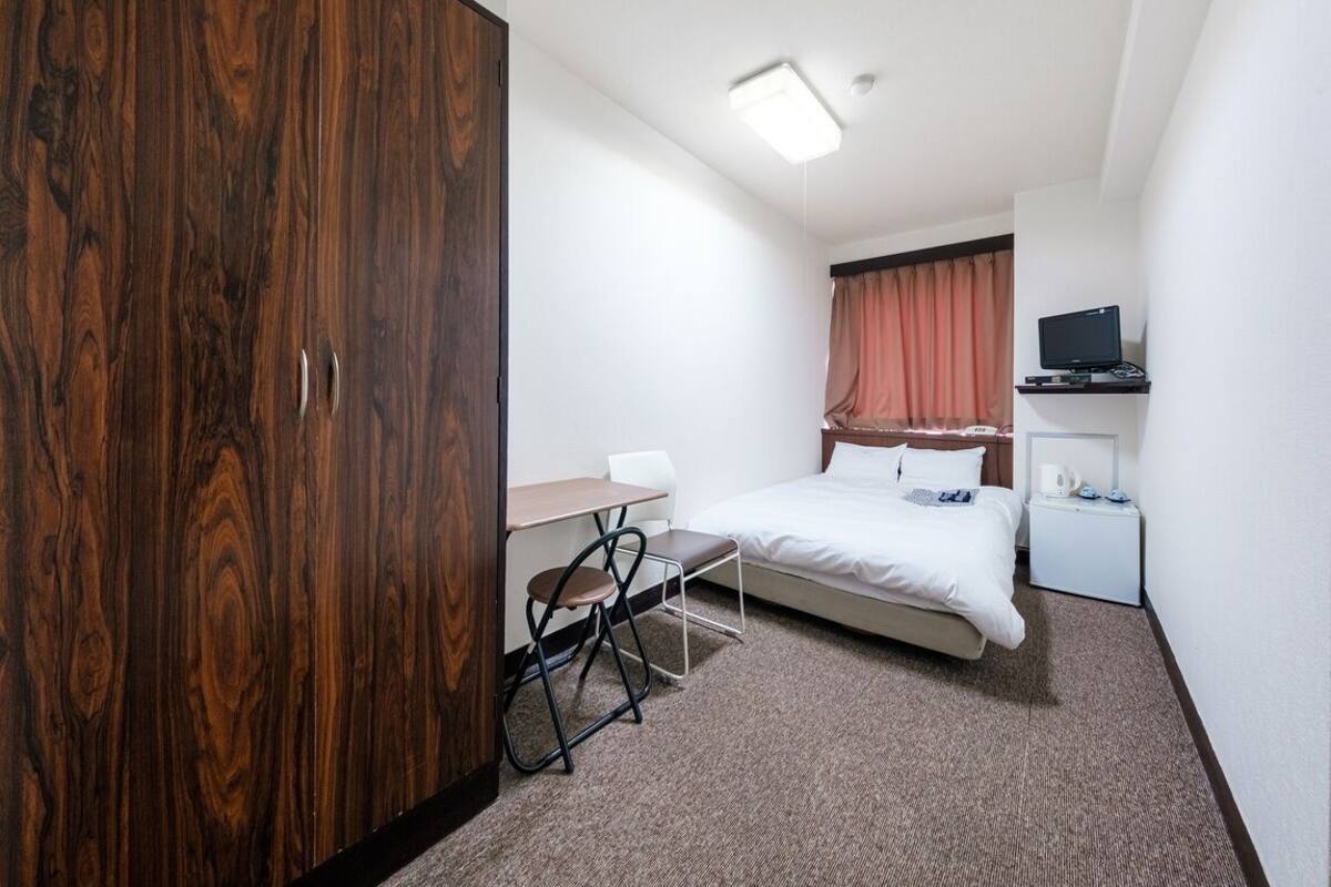 Une chambre d’hôtel de taille réduite, comprenant un lit simple, un bureau avec une chaise, une armoire, un téléviseur posé sur un meuble et une petite table de chevet.