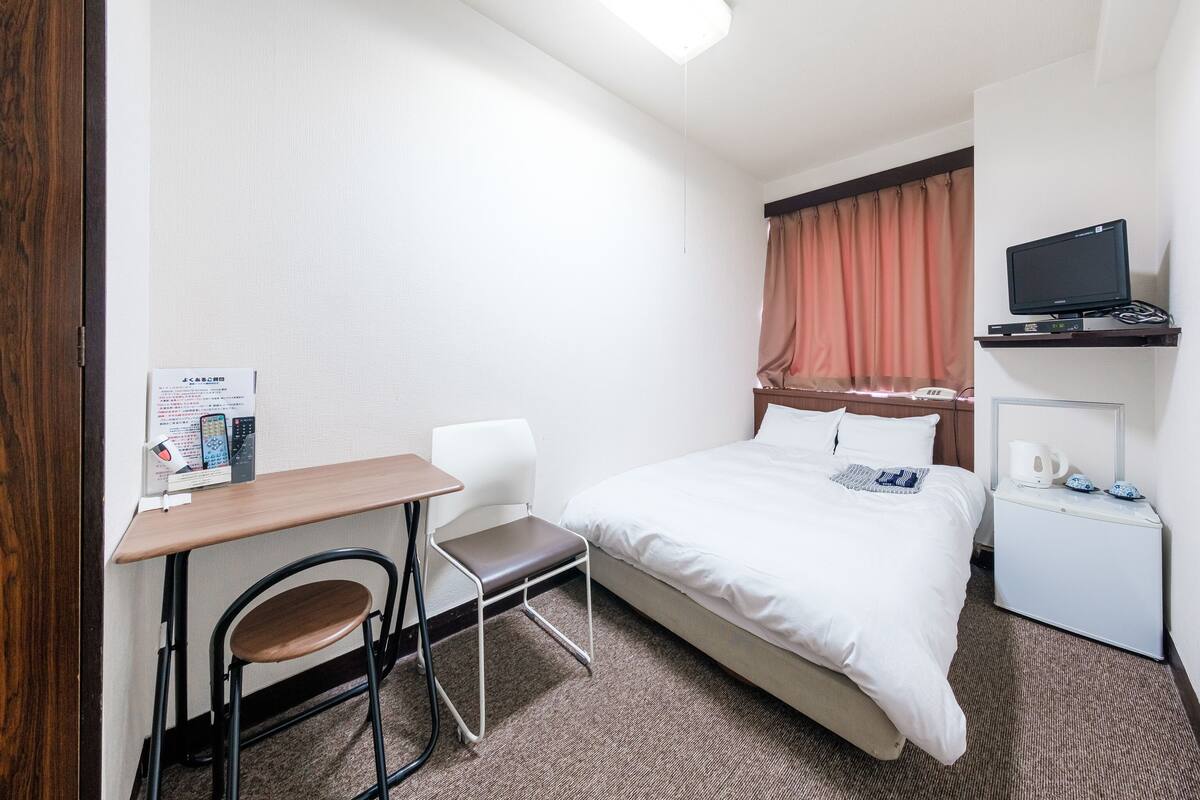 Une chambre d’hôtel avec un lit, un bureau et une chaise, un téléviseur posé sur une étagère et une petite armoire blanche.