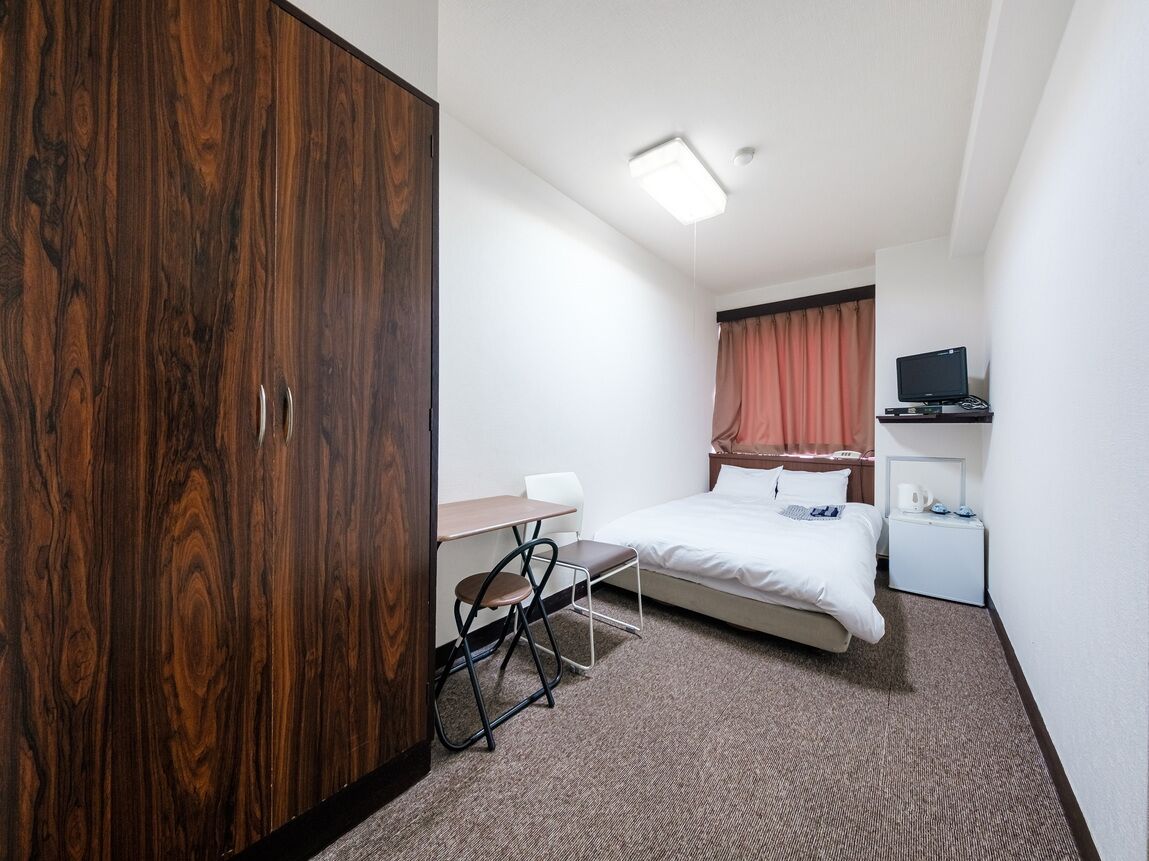 Une chambre d’hôtel de taille réduite, comprenant un lit simple, un bureau avec une chaise, une armoire, un téléviseur posé sur un meuble et une petite table de chevet.
