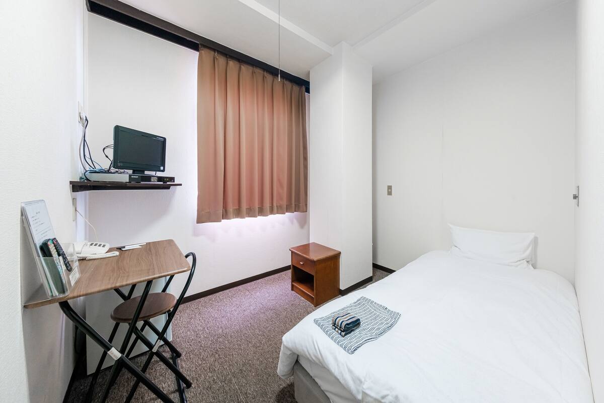 Une chambre d’hôtel avec un lit, un bureau avec un ordinateur, une chaise, une petite table de chevet en bois et une fenêtre avec des rideaux.