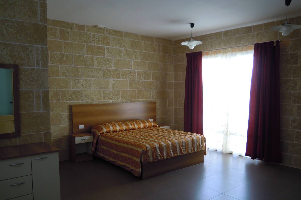 Quarto com uma cama grande, mesa de cabeceira, cômoda e janela com cortinas.