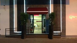 A modern building entrance with a red awning, glass doors, and two potted plants.
