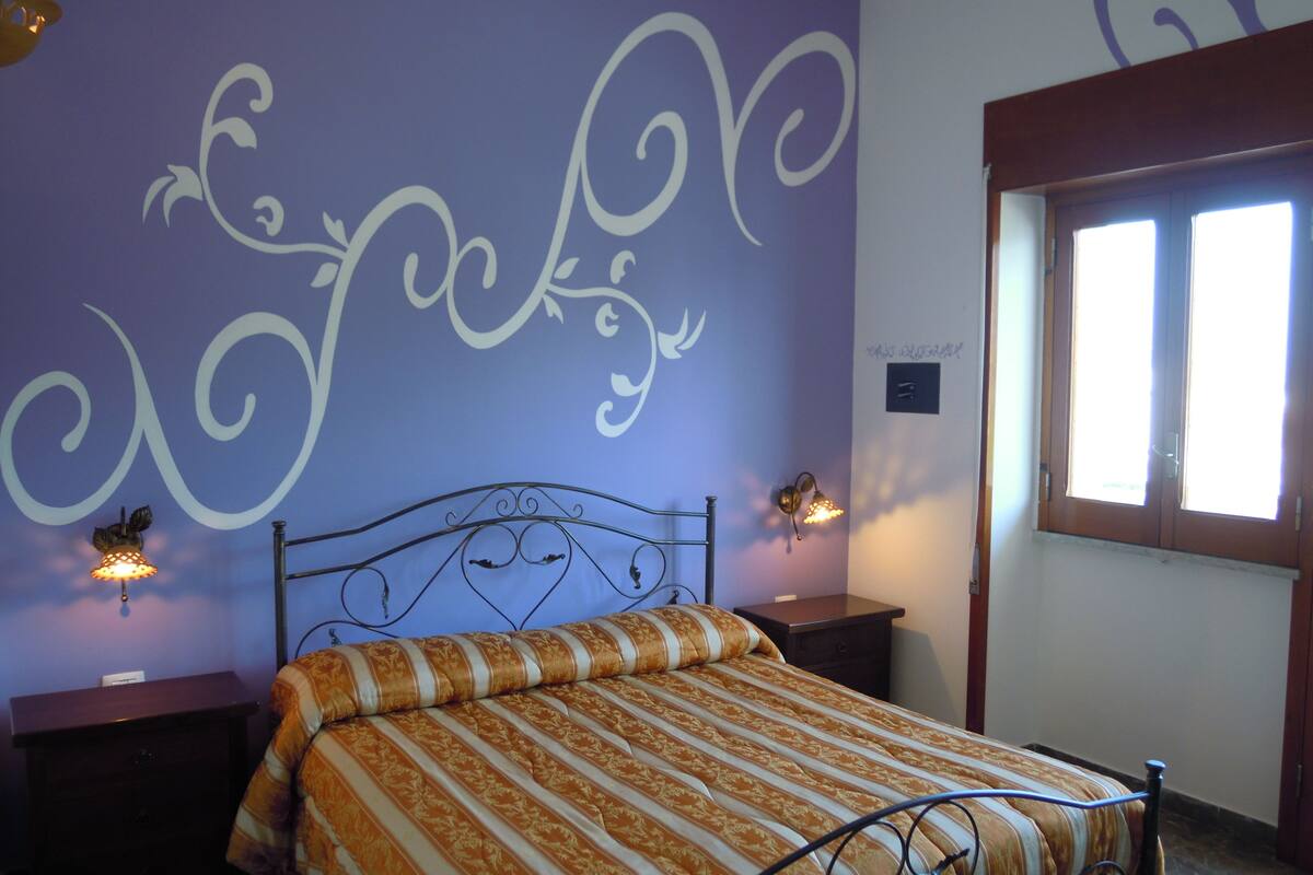 Quarto com cama, duas mesas de cabeceira, parede com detalhes decorativos e uma janela.