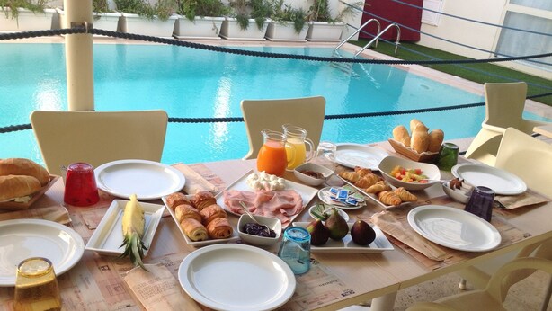 A breakfast table with croissants, fruits, and beverages by a poolside.