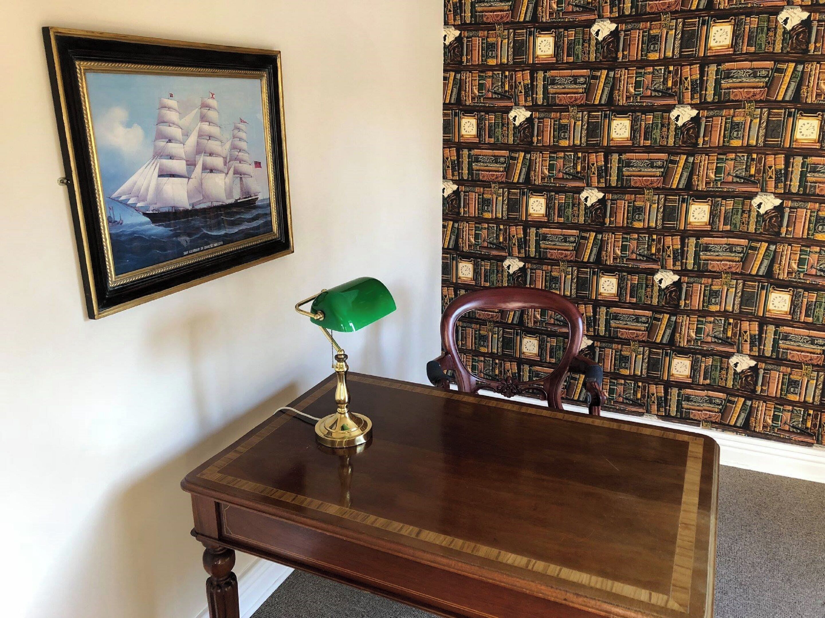 A wooden desk with a green desk lamp, a framed picture of a sailing ship, and a bookshelf with books and clocks.