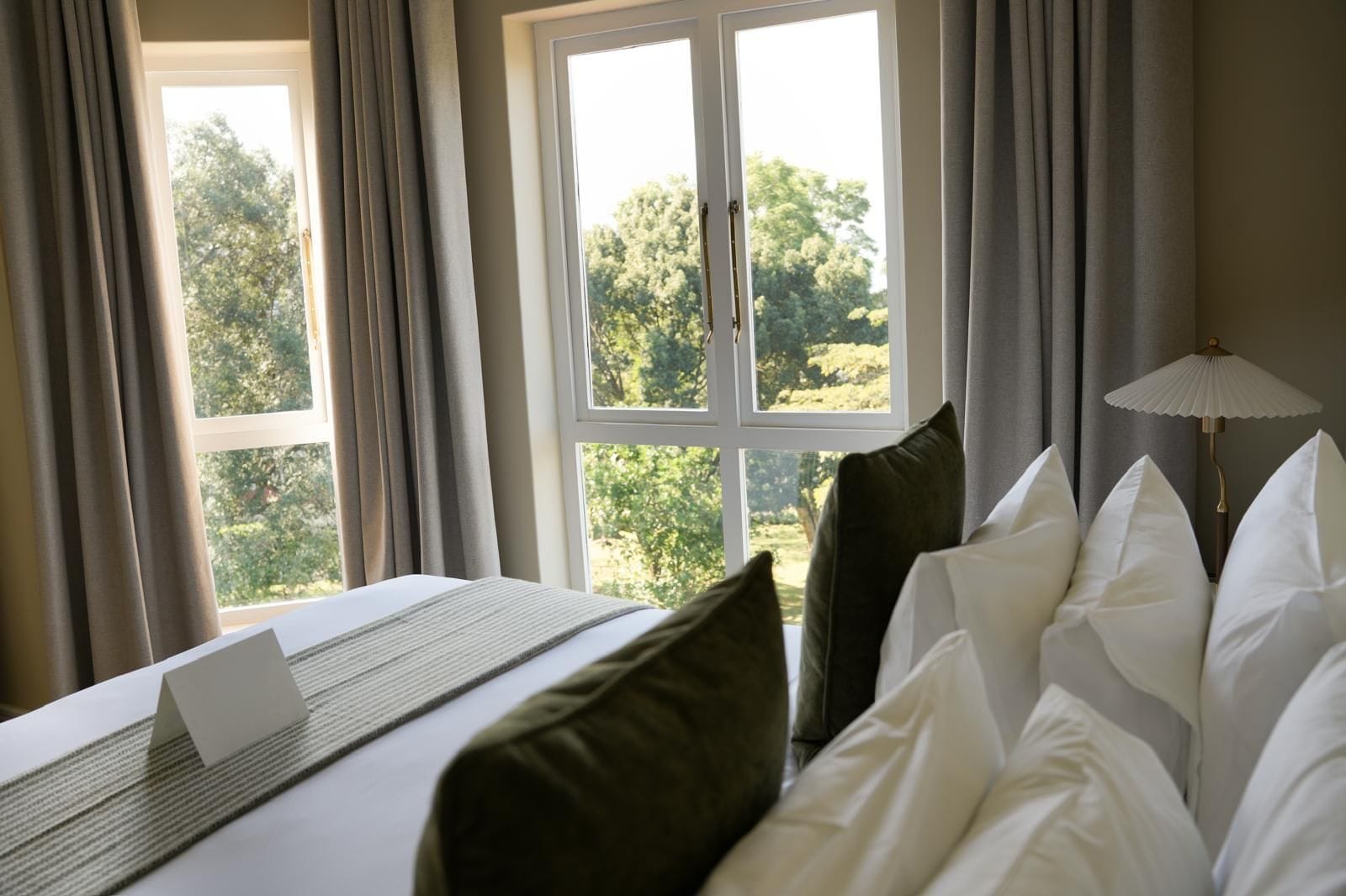 Habitación de hotel con cama, laptop sobre la cama, lámpara y vista a árboles por la ventana.