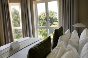 Habitación de hotel con cama, laptop sobre la cama, lámpara y vista a árboles por la ventana.