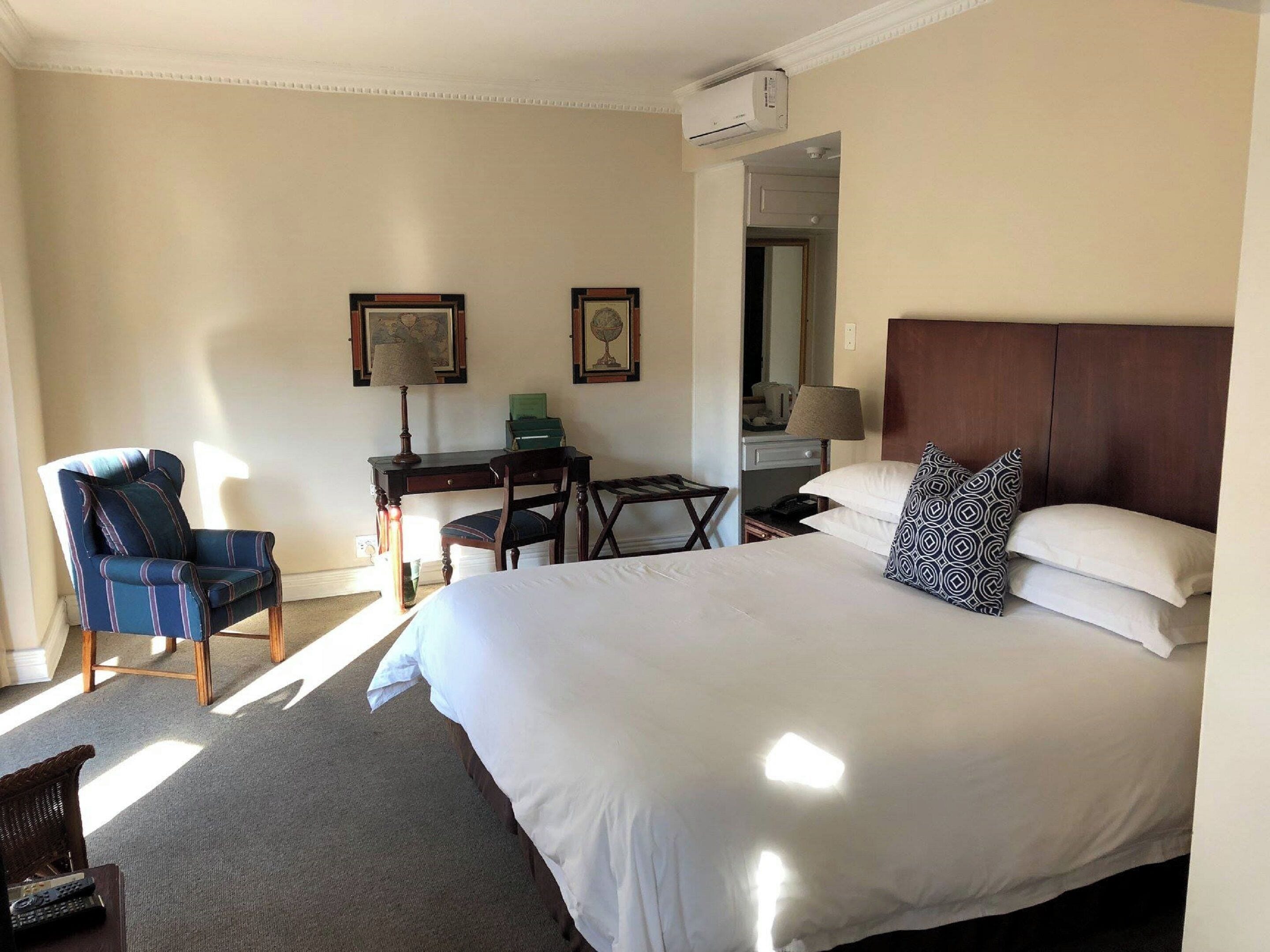 A hotel room with a large bed, a desk, a chair, and a small table. There is a window providing natural light, and an air conditioning unit mounted on the wall.
