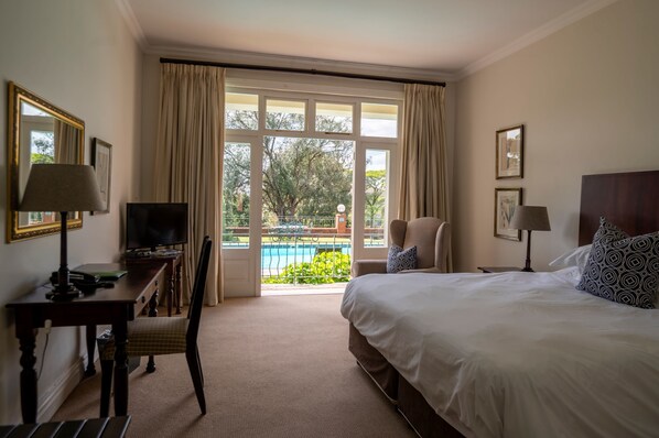 A hotel room with a bed, a desk with a lamp, a chair, a television, and a window with curtains. - Redlands Hotel (Pietermaritzburg)