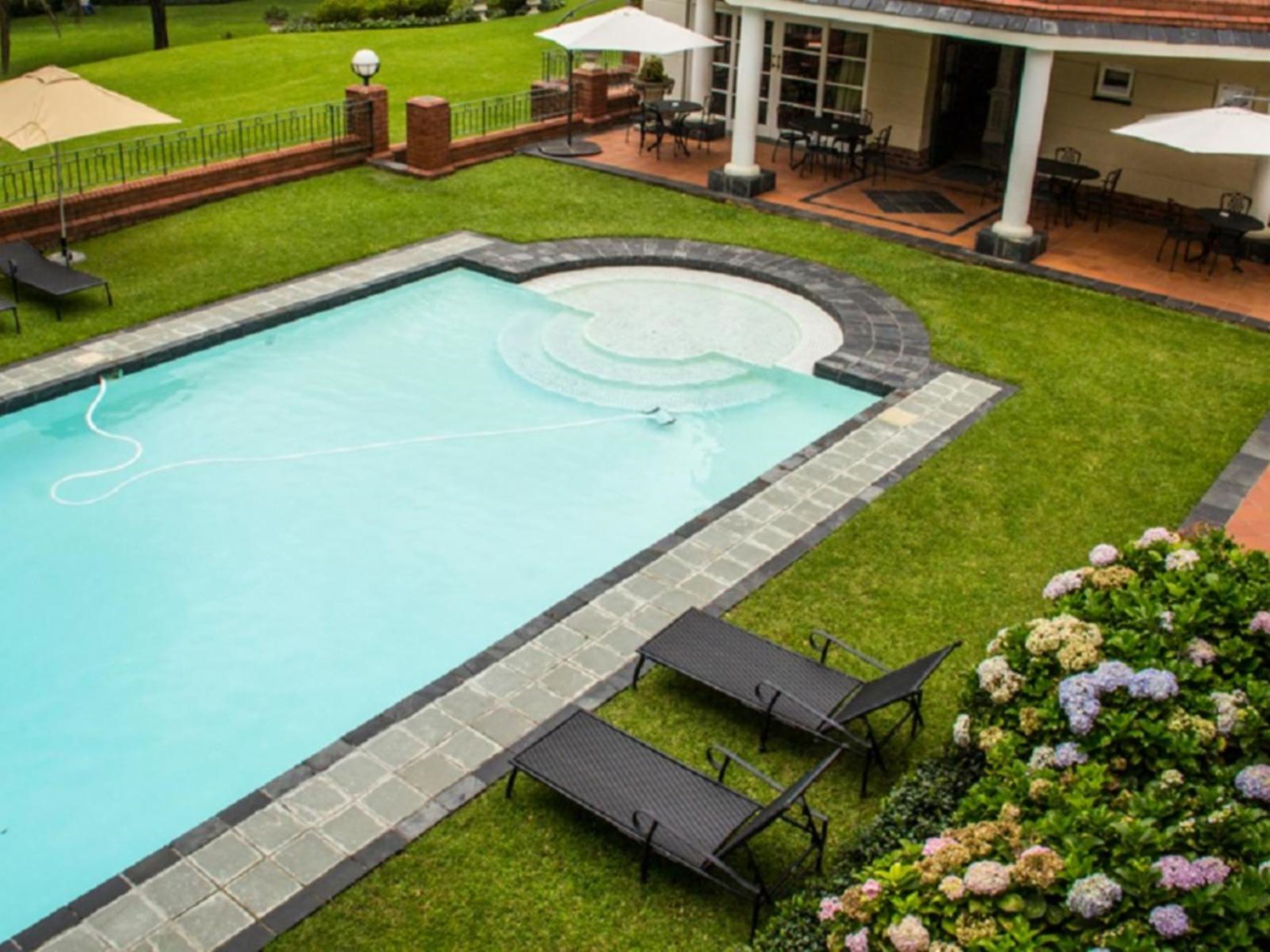 A rectangular swimming pool with a stone border, surrounded by a well-maintained lawn and a patio area with outdoor furniture.