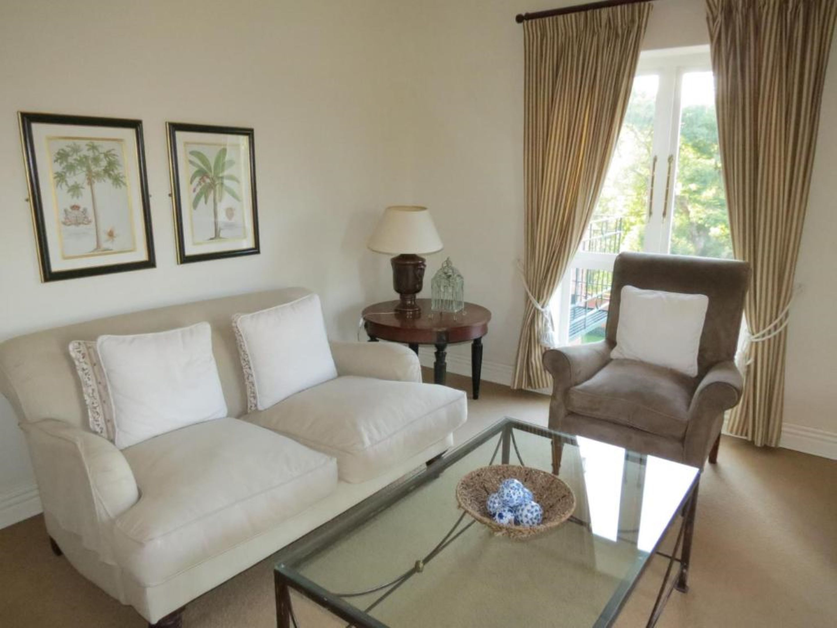 A living room with a beige sofa, a glass coffee table with a decorative bowl, a wooden side table with a lamp, and a brown armchair. There are two framed botanical prints on the wall.