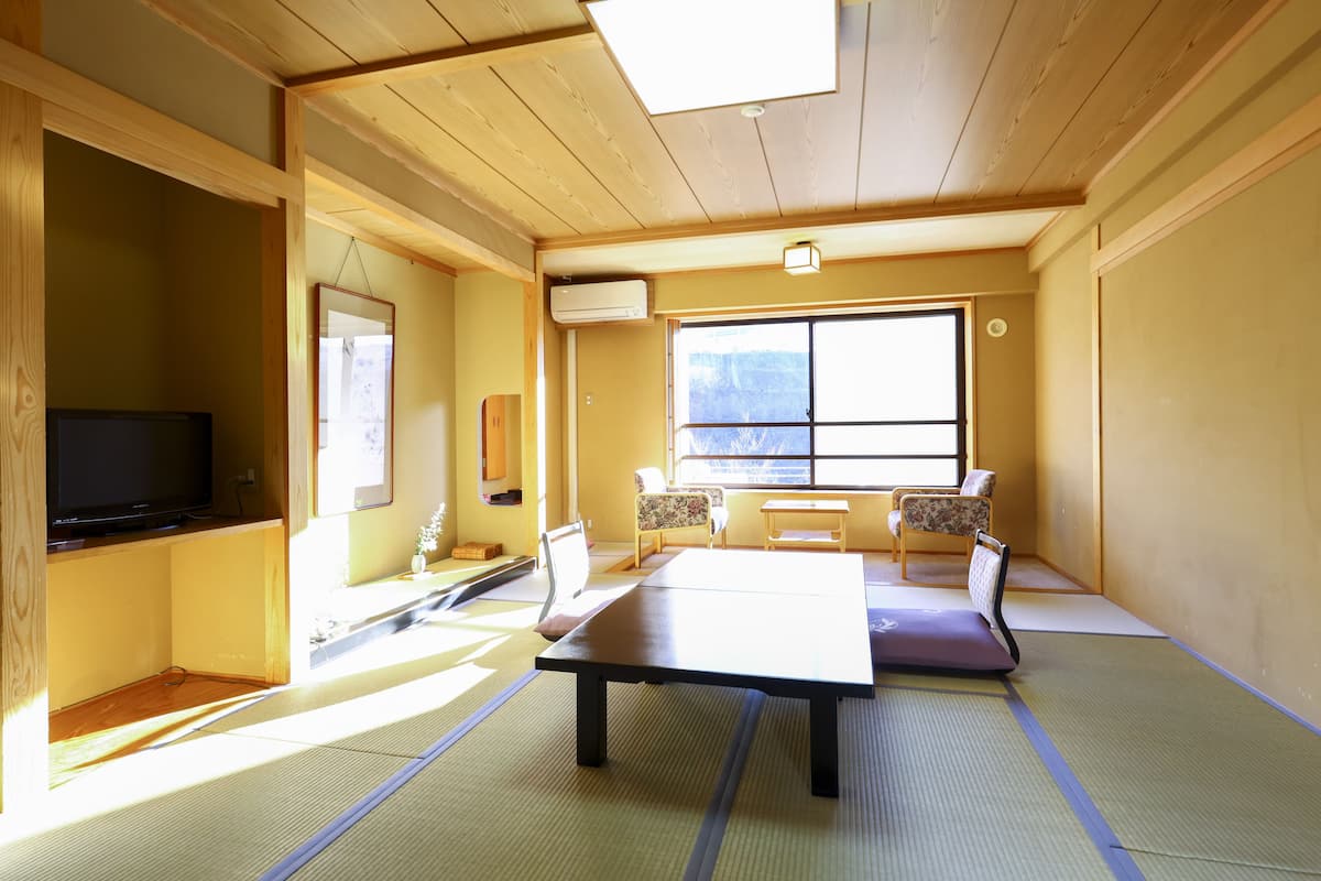 A traditional Japanese room with tatami flooring, a tatami mat table, a television, and a large window with a view of the mountains.