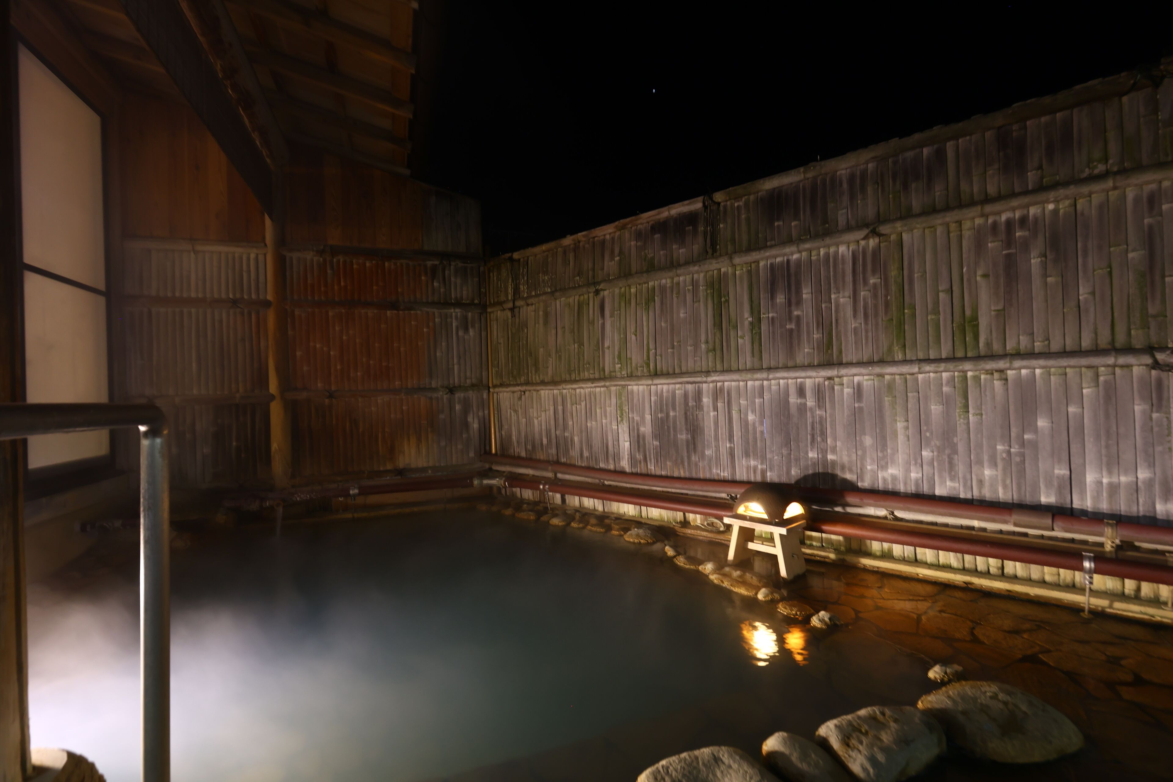 Un onsen japonés tradicional con interior de madera, una fuente termal de piedra y un pequeño banco de madera.