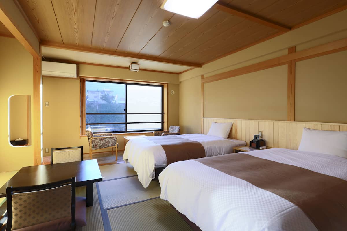 A hotel room with two beds, a small table, a chair, and a window with a view.