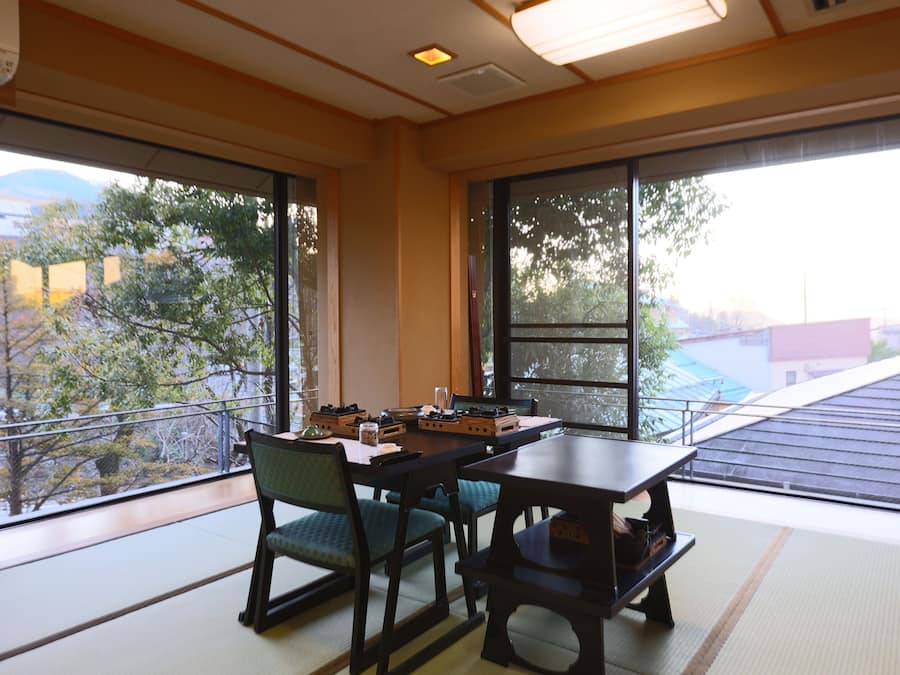 A traditional Japanese room with tatami flooring, a low table, and chairs. Large windows offer a view of trees and buildings outside.