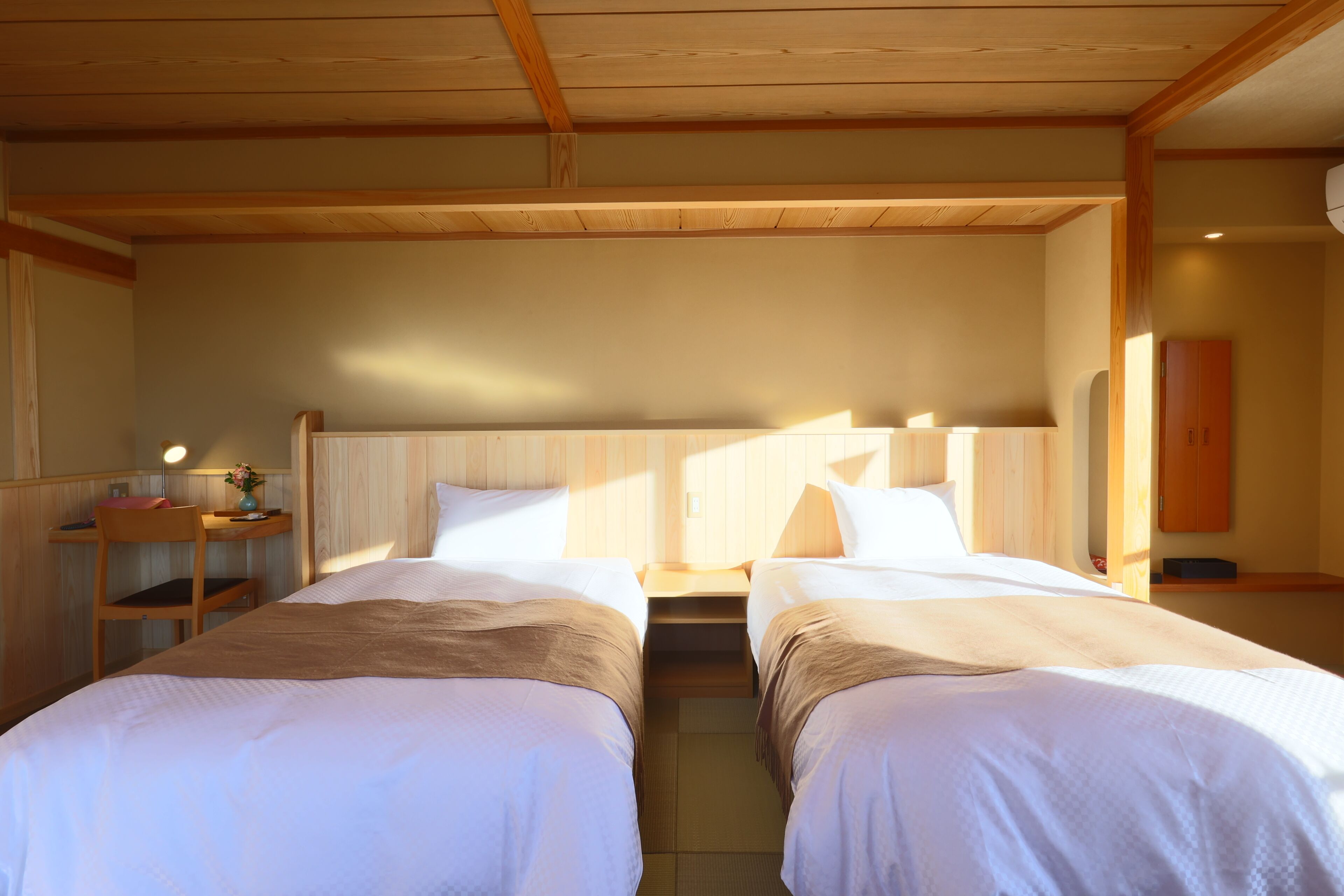 A room with two beds, wooden headboards, a desk with a lamp, and a small table with a vase.