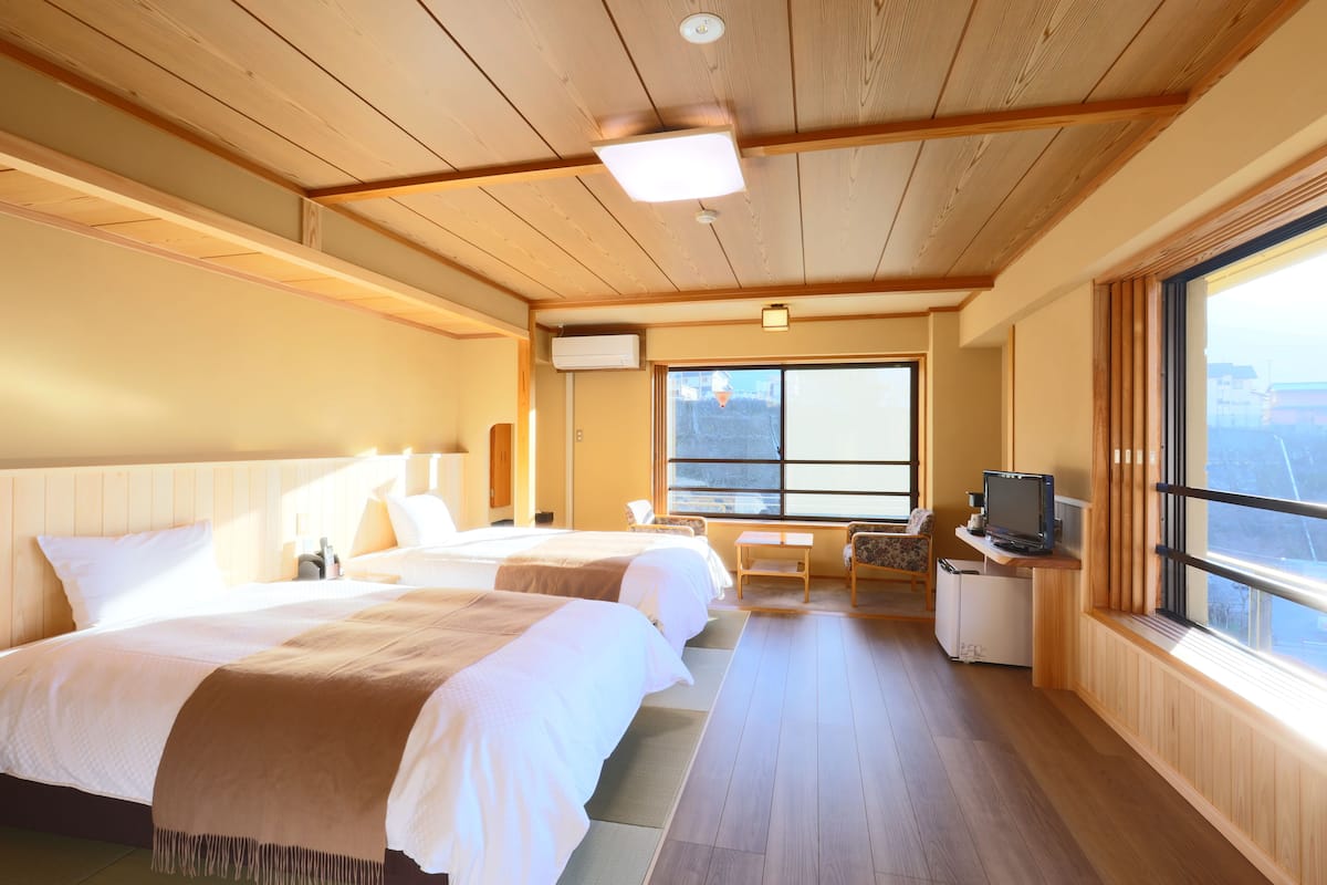 A hotel room with two beds, a desk with a television, a chair, and a large window providing a view.