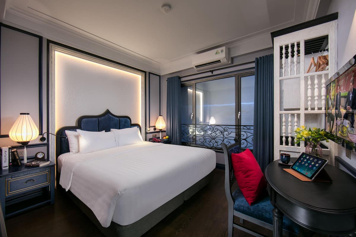 A hotel room with a large bed, bedside tables with lamps, a small table with a tablet, a red pillow, and a view of the outside.