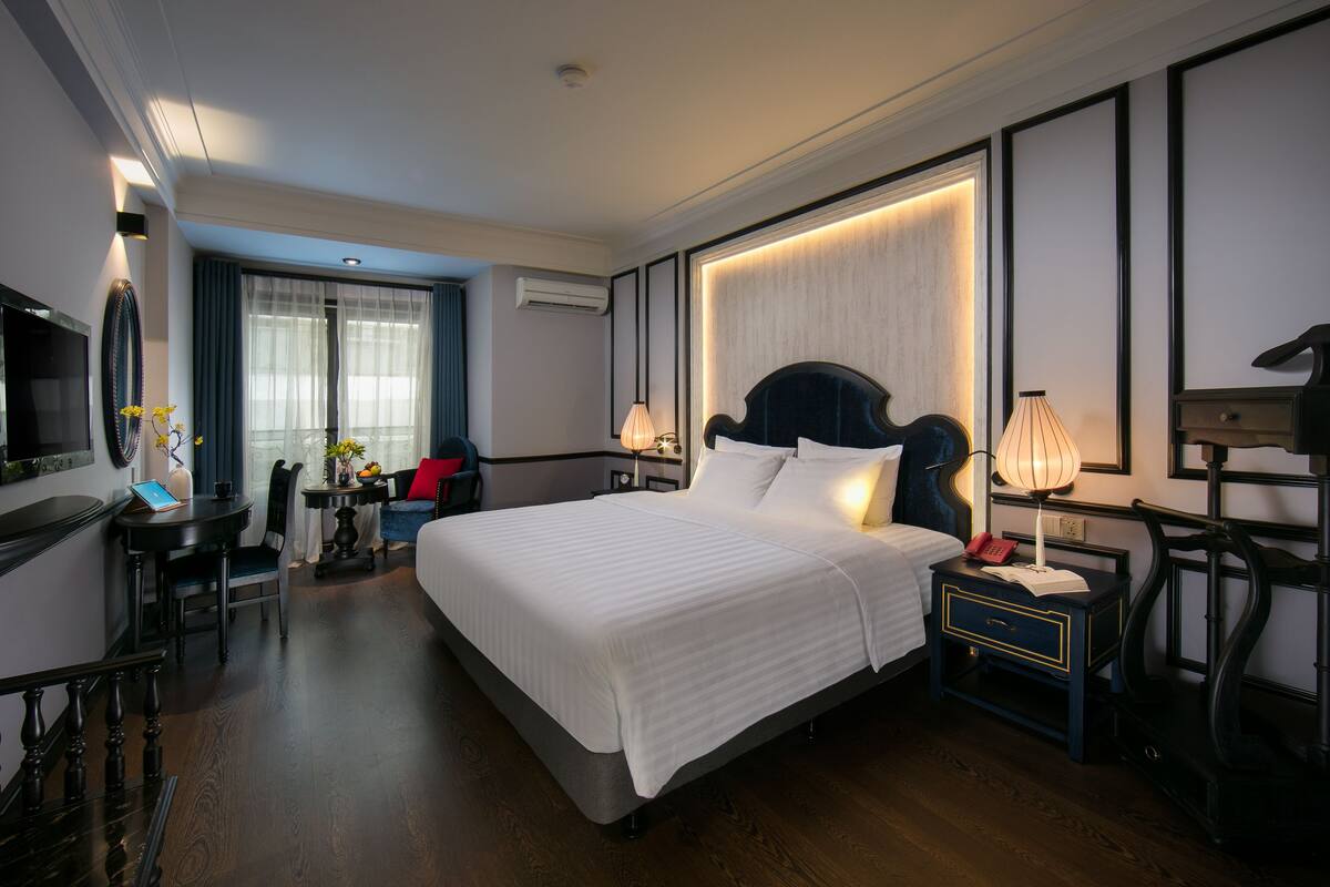 A hotel room with a large bed, bedside tables, a desk with a chair, a TV, and a window with curtains.