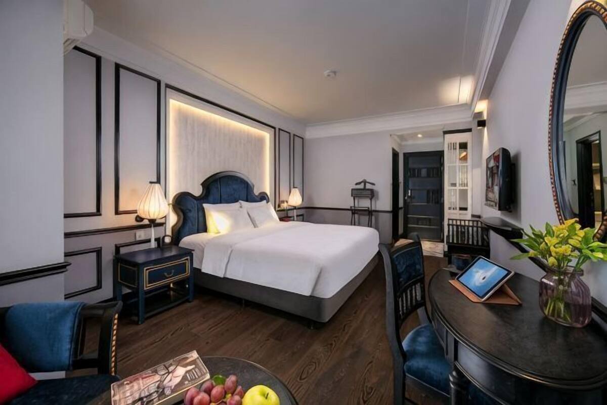 A hotel room with a bed, bedside tables, a desk with a chair, a mirror, and a small table with fruit.