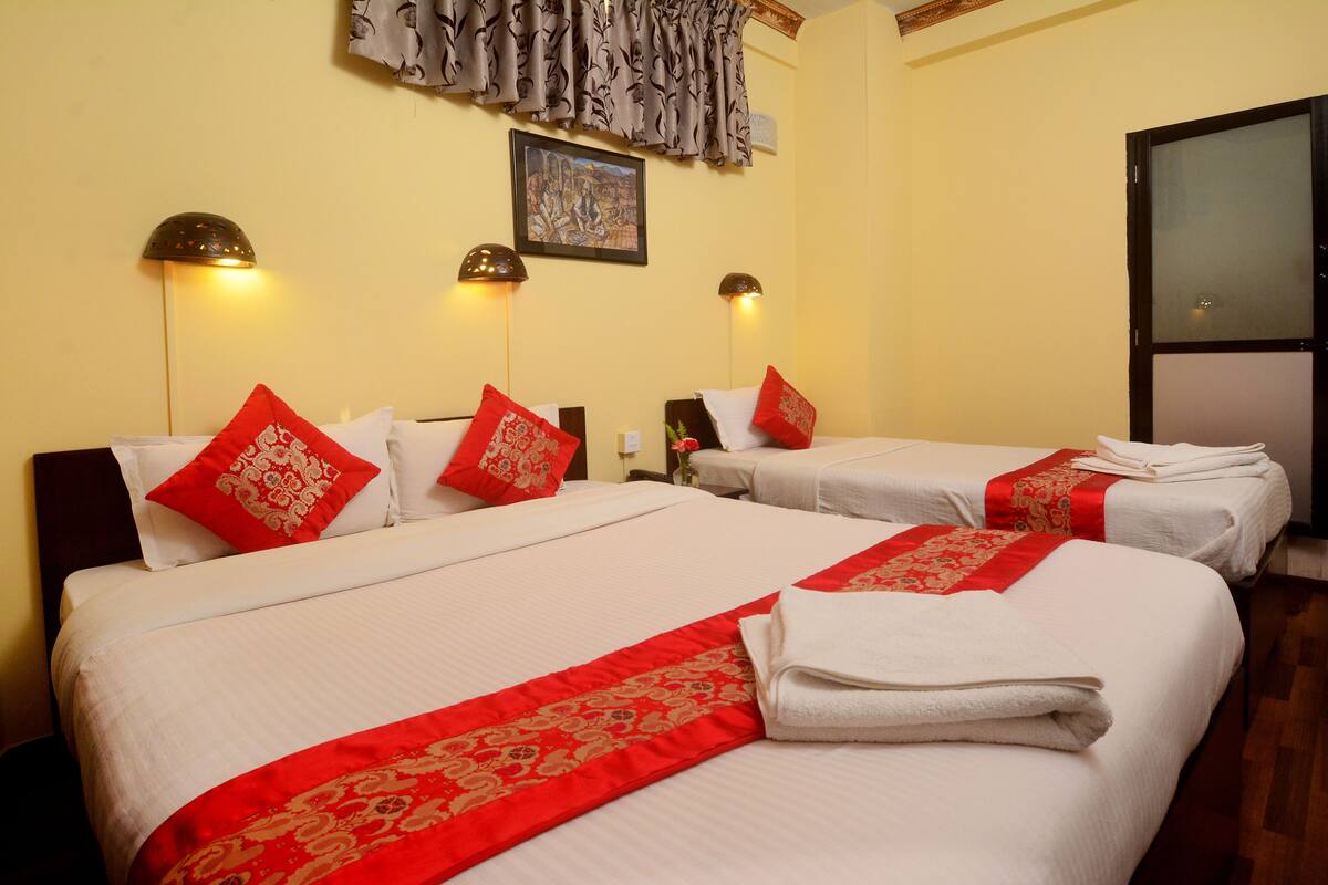 Une chambre d’hôtel avec deux lits, chacun agrémenté de draps blancs et de coussins décoratifs rouges. Des lampes murales et un tableau encadré sont fixés au mur au-dessus des lits.