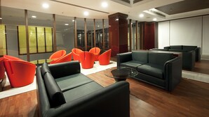A modern lounge area with leather sofas and bright orange chairs, wooden flooring, and large windows with reflections.