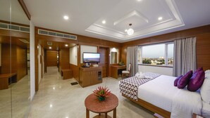 A hotel room with a large bed, a desk with a TV, a small table with a plant, and a view of the city through the window. - JP Hotel (Chennai)