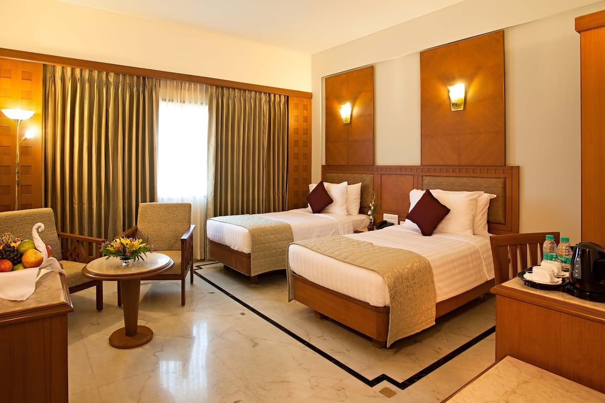 A hotel room with two beds, a desk with a lamp, a chair, a small table with fruit, and a TV.