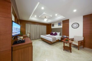 A hotel room with a bed, a desk, a chair, a TV, and a clock on the wall.