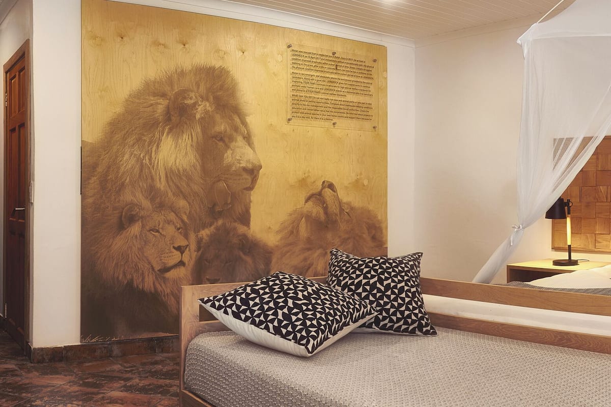 Une chambre à coucher avec un lit à baldaquin, une tête de lit en bois, une table de chevet avec une lampe, une étagère avec de la verrerie et une grande fresque représentant un lion sur le mur.