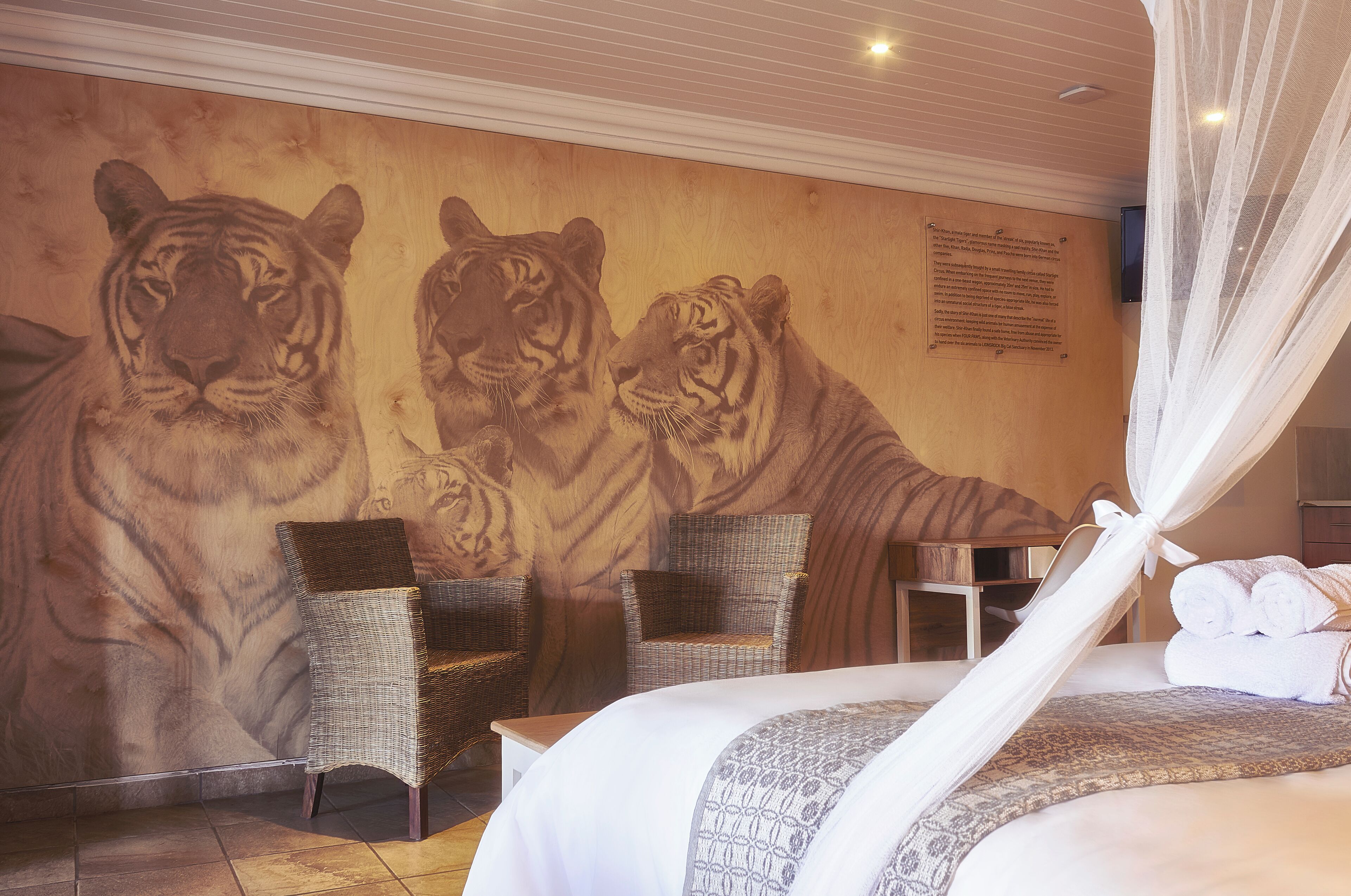Une chambre d'hôtel avec une grande fresque représentant un tigre sur le mur, des meubles en osier et un lit soigneusement fait, surmonté d'un dais.