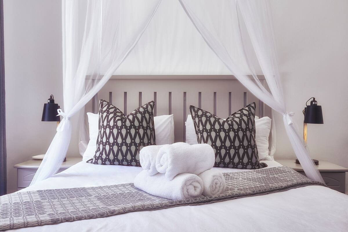 Un lit bien fait, avec des draps blancs, deux coussins à motifs et un baldaquin.