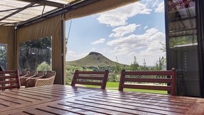 A covered outdoor seating area with a table and chairs, overlooking a grassy landscape and a hill.