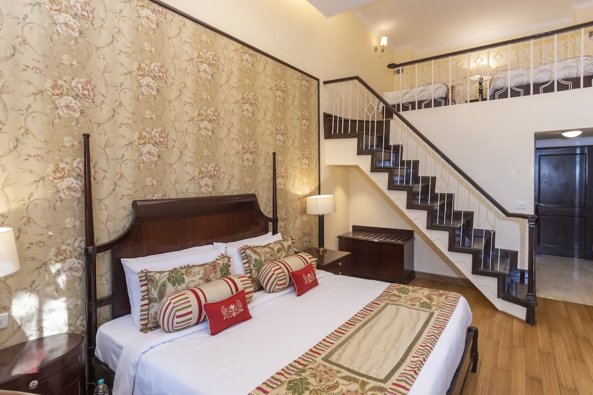 Un dormitorio con una cama grande, cabecera de madera, papel tapiz floral, una mesita de noche con lámpara y una escalera que conduce a un piso superior.