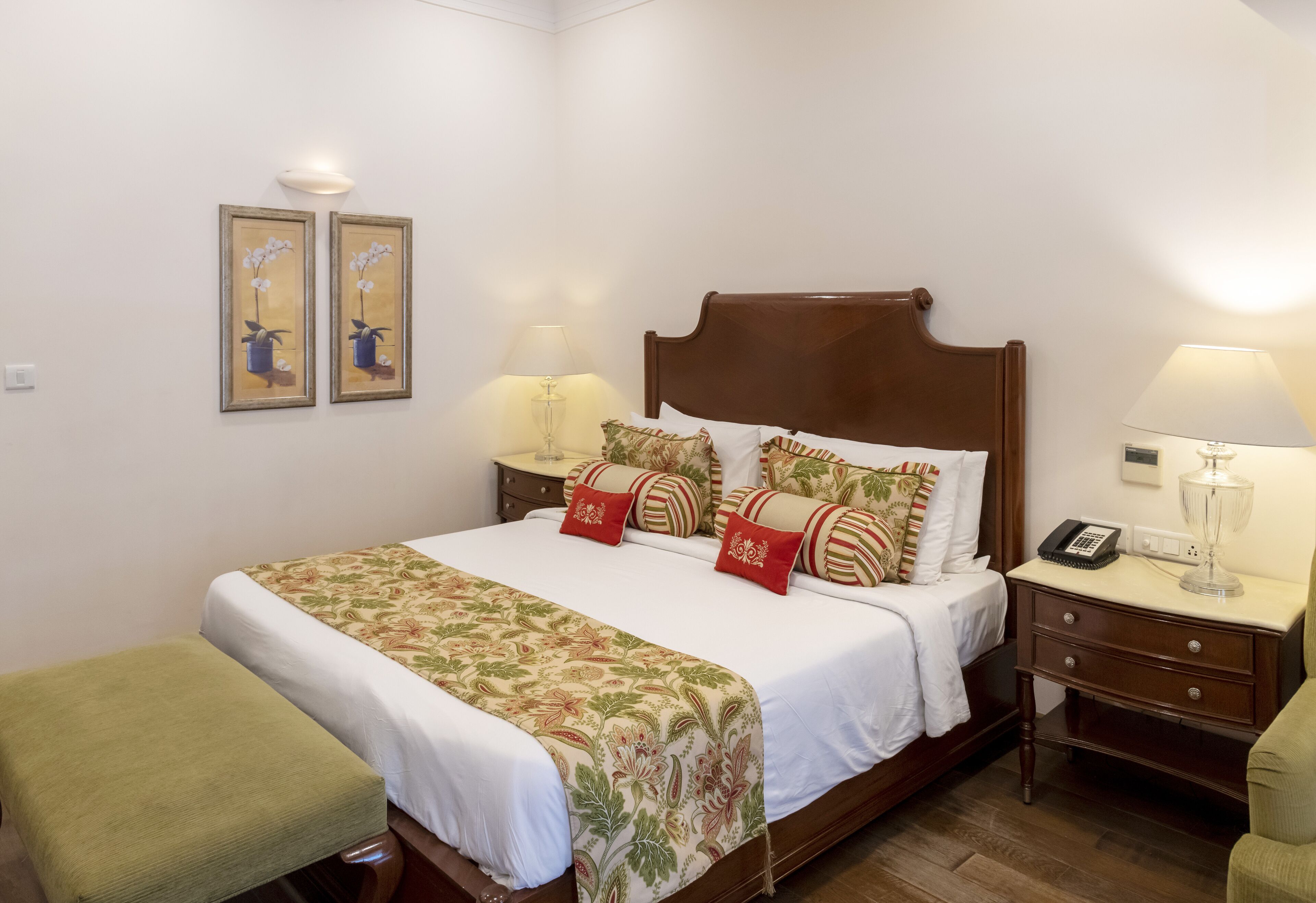 Una cama bien hecha con una colcha estampada con motivos florales, dos lámparas de noche, un teléfono junto a la cama y un pequeño banco verde.