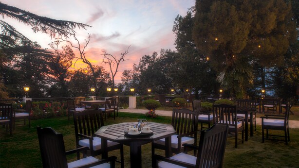 Un área de comedor al aire libre con mesas y sillas redondas, rodeada de árboles y con un atardecer de fondo.