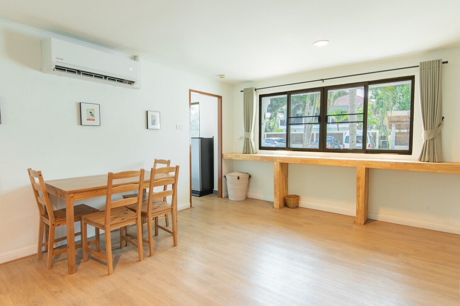 A room with a wooden dining table and chairs, a window with curtains, and a trash bin.