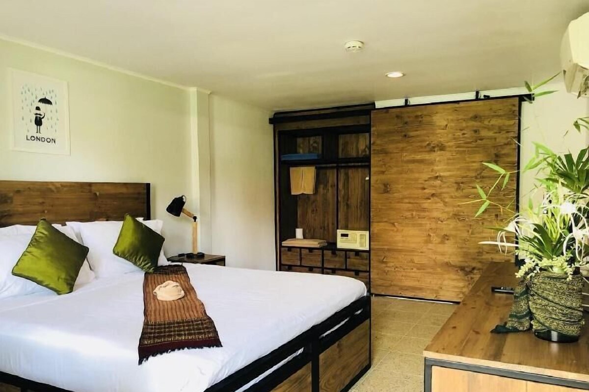 A bedroom with a bed, pillows, a bedside table, a lamp, a wooden wardrobe, and a plant.