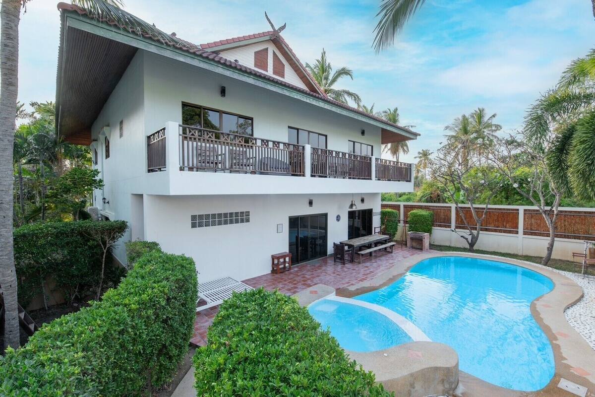A two-story house with a swimming pool, surrounded by lush greenery and palm trees.