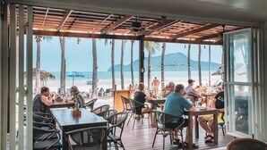 An outdoor dining area with tables and chairs, overlooking a beach with palm trees and a mountain in the distance. - Dolphin Bay Beach Resort (Sam Roi Yot)