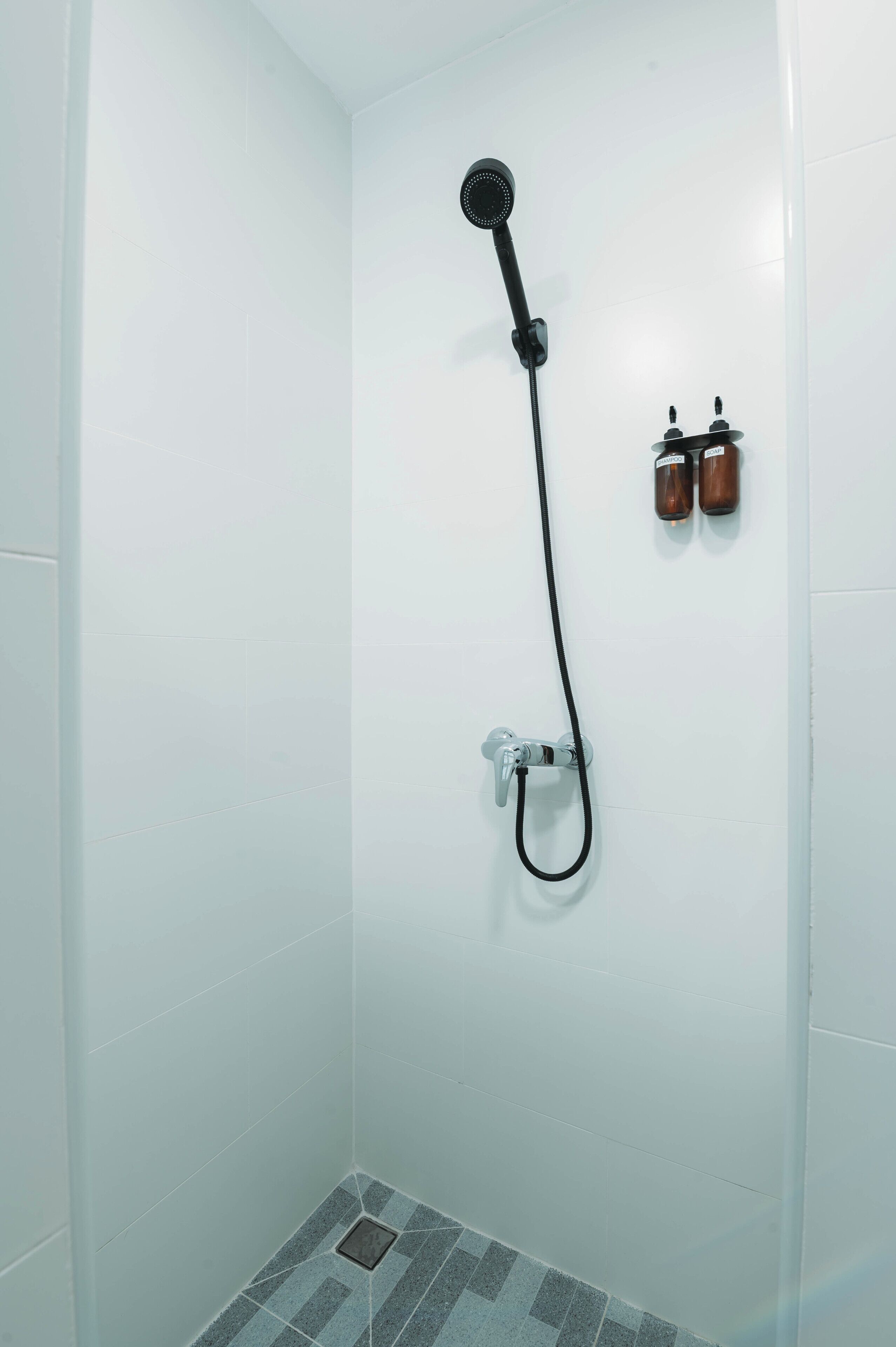 Un baño pequeño con azulejos blancos y un cabezal de ducha de mano, con dos botellas de champú en la pared.