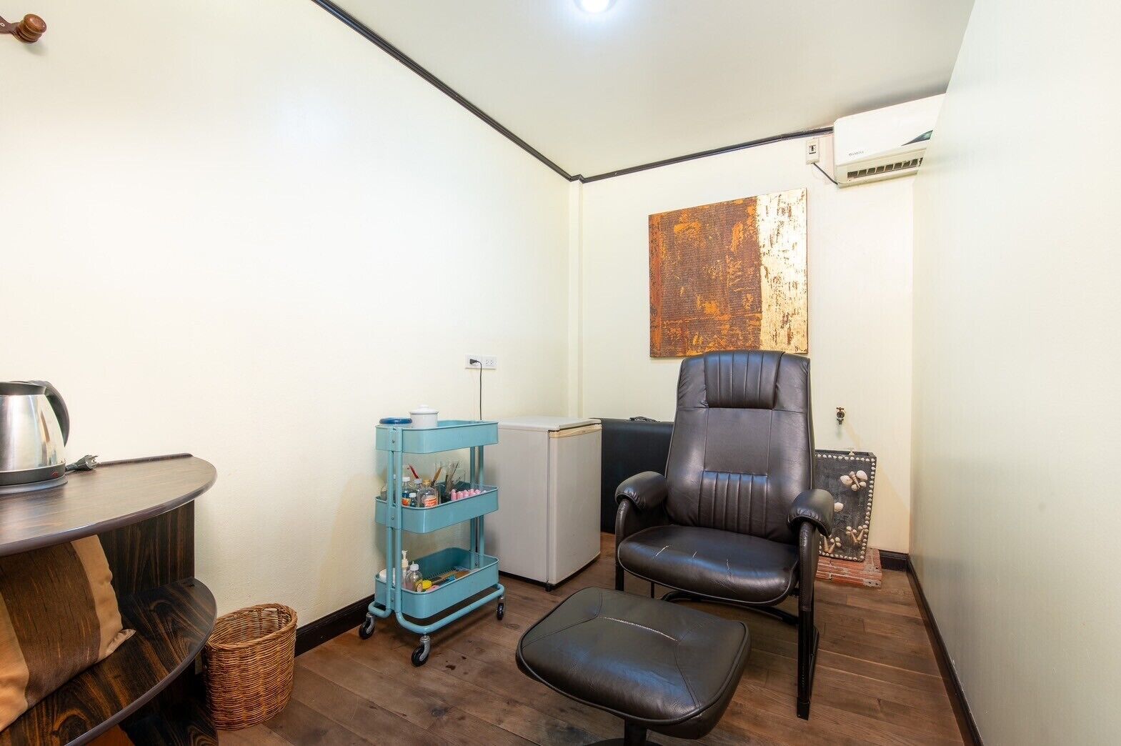 Una habitación pequeña y funcional con suelo de madera, una silla de cuero negro, una mesita con una tetera, un carrito azul con productos de limpieza y un radiador.