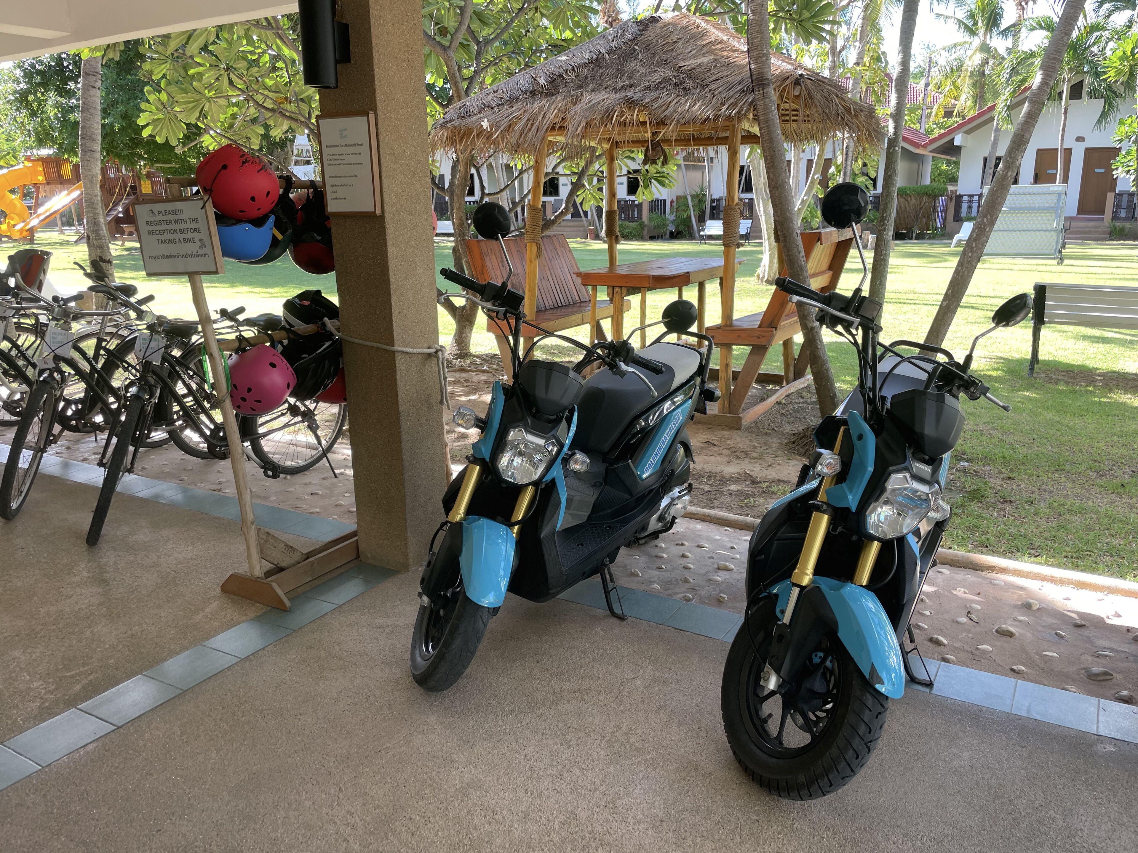 A covered parking area with two blue scooters, helmets, and a sign indicating registration for bike rental.